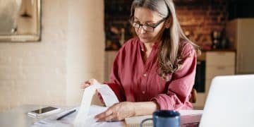 Serious focused middle aged female accountant in eyewear working from home providing outsource bookkeeping service, sitting at table with portable computer, calculating finances, doing paperwork